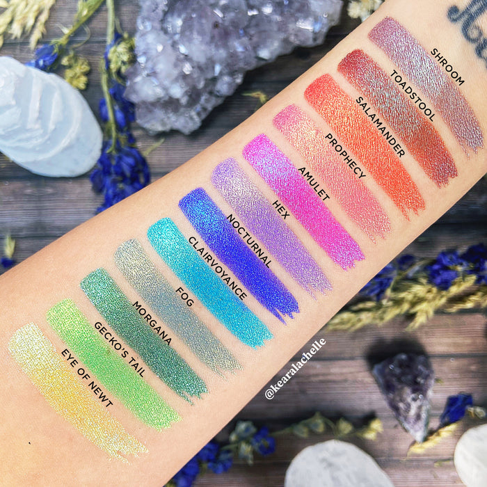 Top angled arm swatches on fair skin tone of Witchcraft vs. Alchemy Collection including Salamander Duochrome Eyeshadow next to Shroom, Toadstool, Fog, Prophecy, Hex, Nocturnal, Clairvoyance, Amulet, Morgana, Gecko's Tail, Eye of Newt