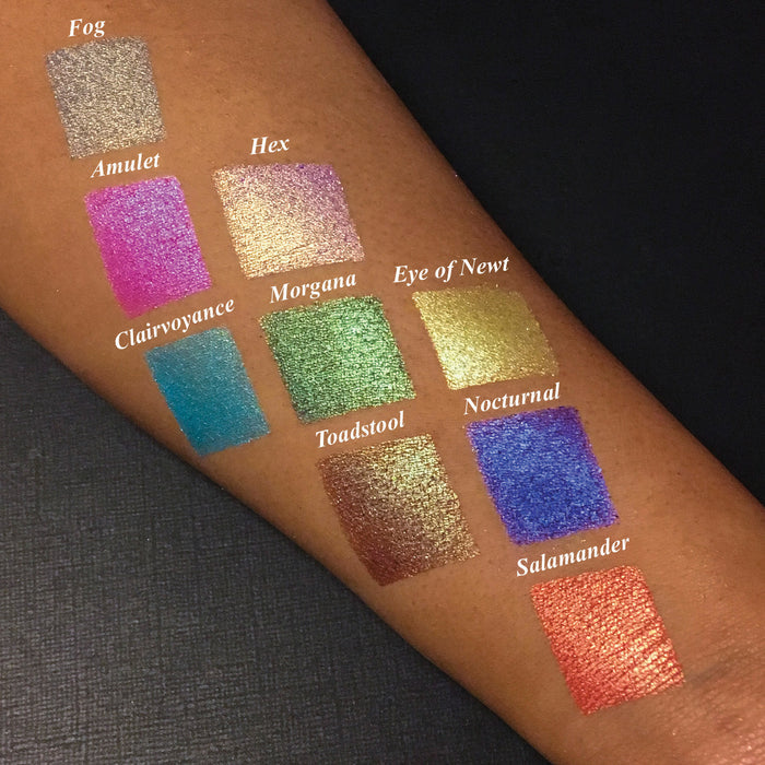 Top angled arm swatches on medium skin tone of Salamander Duochrome Eyeshadow next to Amulet, Hex, Clairvoyance, Morgana, Eye of Newt, Toadstool, Nocturnal, Fog