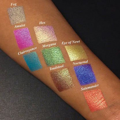 Top angled arm swatches on medium skin tone of Salamander Duochrome Eyeshadow next to Amulet, Hex, Clairvoyance, Morgana, Eye of Newt, Toadstool, Nocturnal, Fog