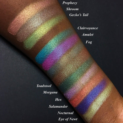 Top angled arm swatches on deep skin tone of Witchcraft vs. Alchemy Collection including Salamander Duochrome Eyeshadow next to Prophecy, Shroom, Gecko's Tail, Clairvoyance, Amulet, Toadstool, Morgana, Hex, Nocturnal, Eye of Newt, Fog