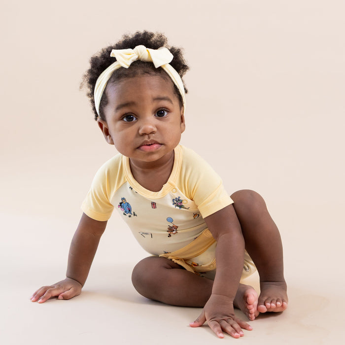 Young toddler sitting on the the floor wearing the Shortall in Winnie the Pooh Celebration with a light yellow bow