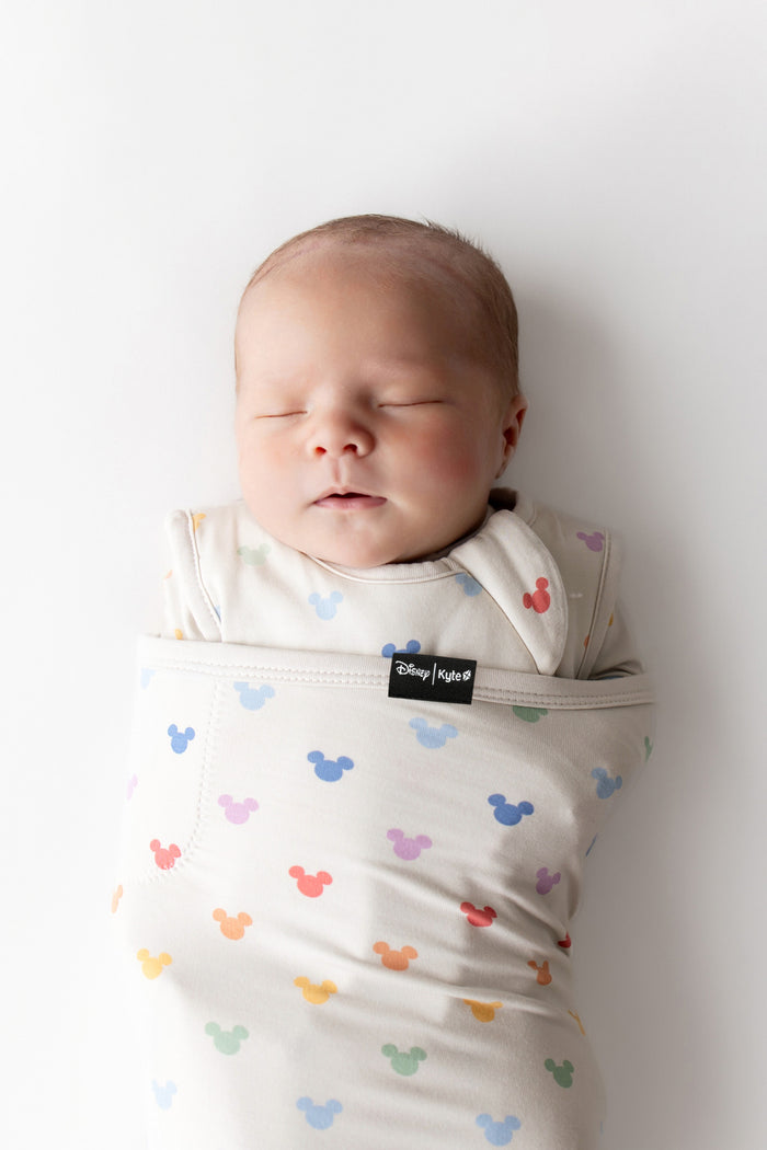 Newborn baby swaddled in a colorful Mickey Mouse patterned swaddle blanket against a white background