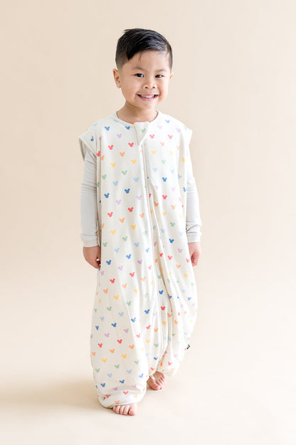 Child wearing a cream sleeveless sleep bag walker with colorful mickey patterns on a beige background