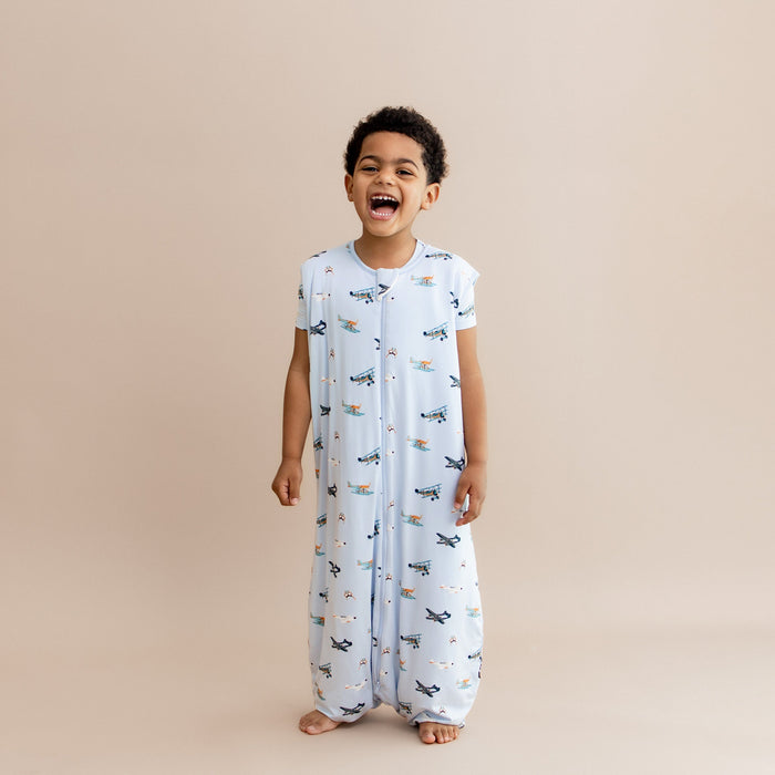 Laughing boy standing wearing the Sleep Bag Walker in Take Off 1.0 with matching short sleeve toddler pajamas on underneath
