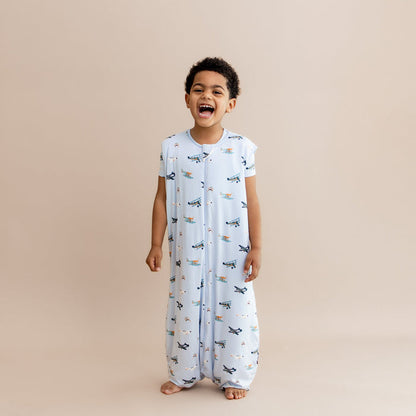 Laughing boy standing wearing the Sleep Bag Walker in Take Off 1.0 with matching short sleeve toddler pajamas on underneath