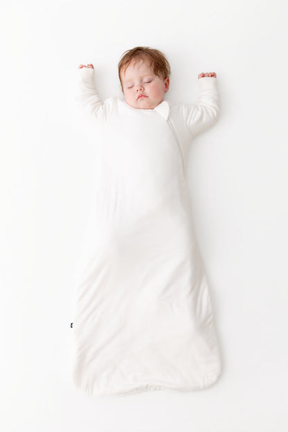 A sleeping newborn baby wearin a white long-sleeve sleep sack, lying on a clean white background.