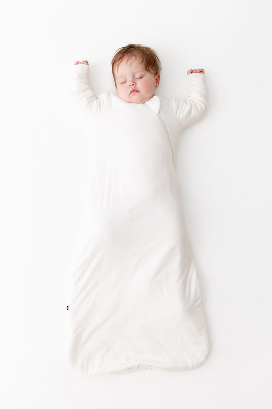 A sleeping newborn baby wearin a white long-sleeve sleep sack, lying on a clean white background.