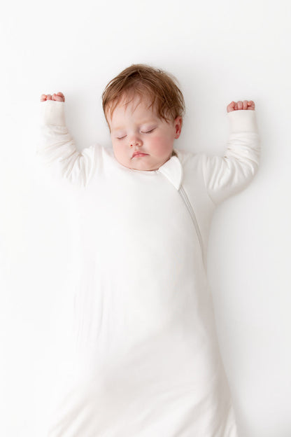 A sleeping newborn baby wearing a long-sleeve white sleep sack, lying flat on a clean white background with arms raised.