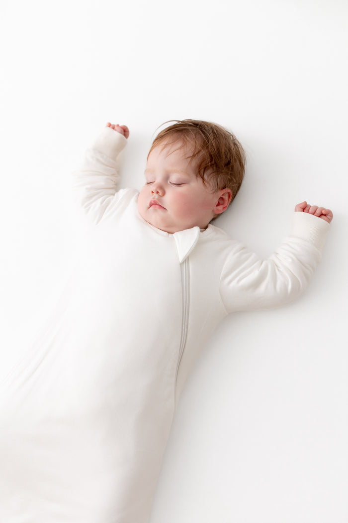A sleeping baby lying on their back wearing a white long-sleeve zip-up sleeper against a clean white background.