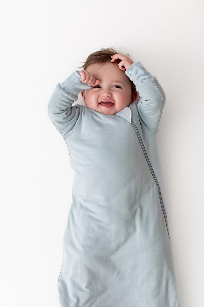 A baby wearing a light blue long-sleeve sleep sack, lying on a plain white background with their hands near their face.