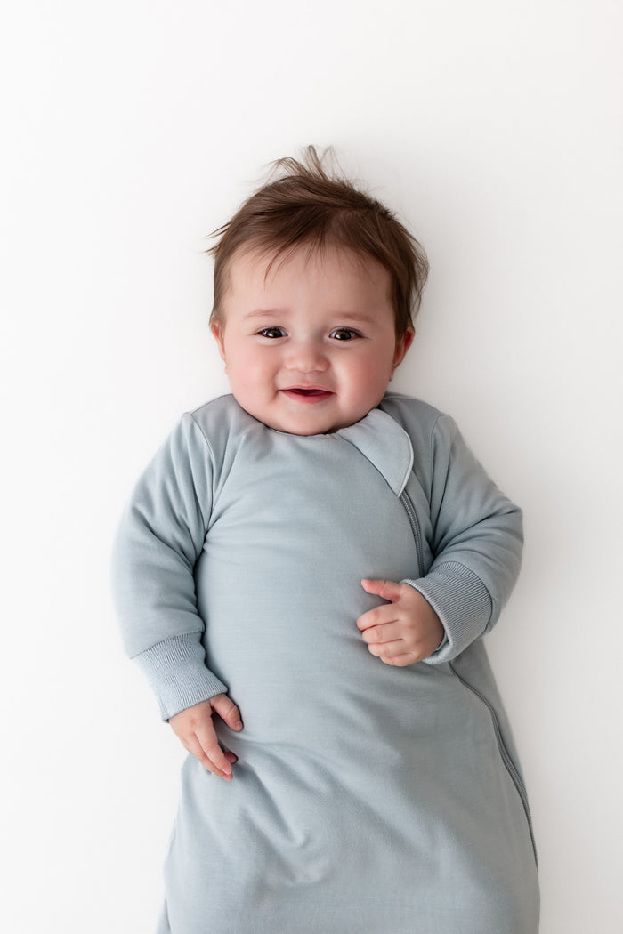 A baby lying on their back wearing a light blue long-sleeve sleep sack against a plain white background.