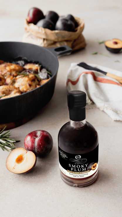 Bottle of Smoky Plum syrup on a table with plums and a skillet in the background