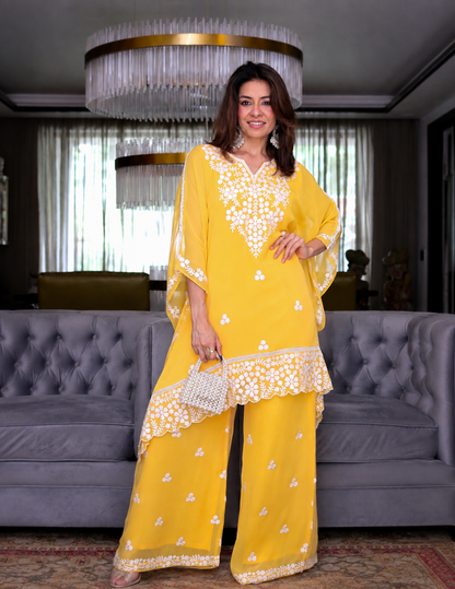 A woman models a bright yellow traditional outfit with white floral embroidery, standing in a modern living room setting with a gray tufted sofa and a large hanging chandelier in the background.