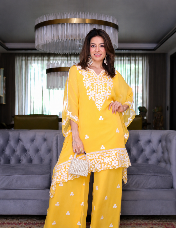 A woman modeling a bright yellow traditional outfit with white floral embroidery, standing in a modern living room with a gray tufted sofa and a large hanging chandelier behind her.
