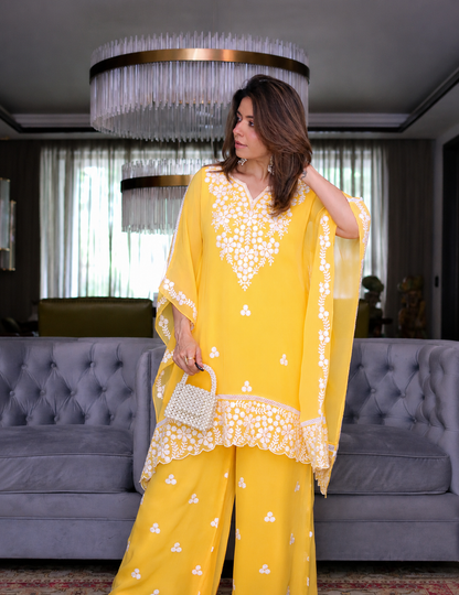 A woman models a bright yellow three-piece traditional outfit with intricate white floral embroidery, holding a small white woven handbag in a modern living room setting.