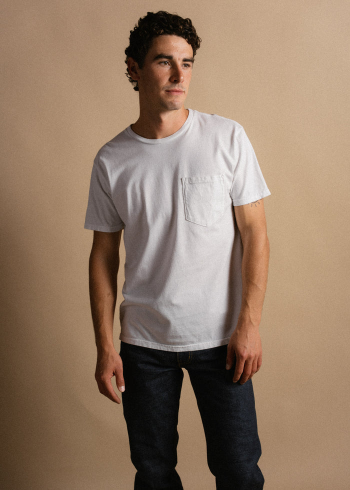 Man in a white T-shirt and jeans stands against a beige background, looking to the side with a thoughtful expression.