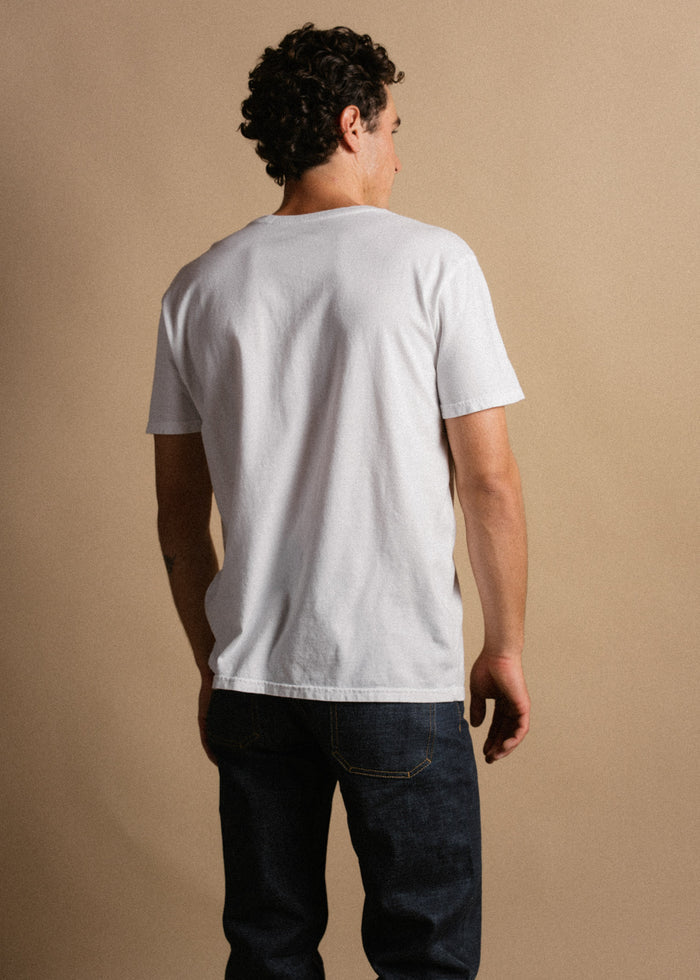 Man in a white t-shirt and jeans stands with his back to the camera against a beige background.