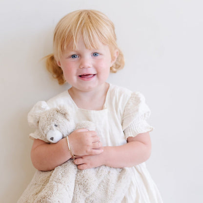 Adorable toddler girl in a white dress cuddling a Saranoni bear stuffed animal lovey. This ultra-soft plush lovey blanket features a cozy bear head and luxuriously soft fabric, designed to provide comfort, security, and soothing snuggles for babies and toddlers. Made with high-quality materials, the Saranoni lovey is perfect for newborns, infants, and young children as a bedtime companion or travel comfort item. Ideal baby shower gift or nursery essential, offering warmth and softness in a neutr