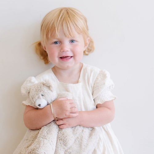 Adorable toddler girl in a white dress cuddling a Saranoni bear stuffed animal lovey. This ultra-soft plush lovey blanket features a cozy bear head and luxuriously soft fabric, designed to provide comfort, security, and soothing snuggles for babies and toddlers. Made with high-quality materials, the Saranoni lovey is perfect for newborns, infants, and young children as a bedtime companion or travel comfort item. Ideal baby shower gift or nursery essential, offering warmth and softness in a neutr