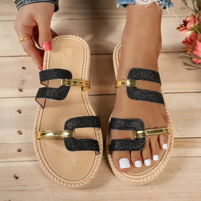 Beige sandals with black and gold accents on a wooden floor.