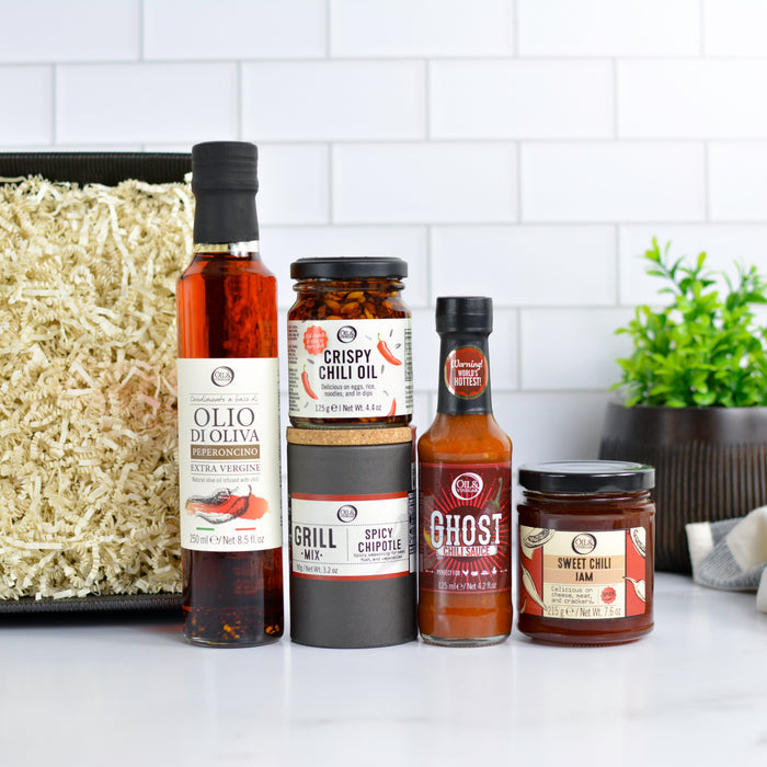 Assorted food products including a bottle of olive oil, a jar of chipotle mix, a jar of sweet chili jam, hot sauce, and chili oil on a kitchen counter.