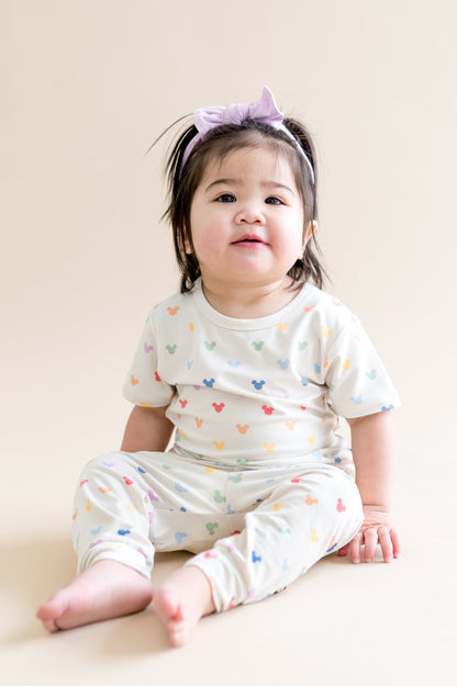 Young toddler wearing a cream tshirt and pant with colorful mickey head patterns, sitting on a beige surface.