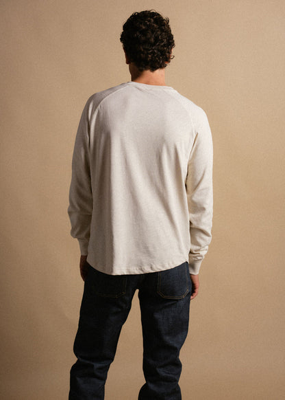 Man in a white long-sleeve shirt and jeans stands with back to camera against a beige background, highlighting casual fashion.