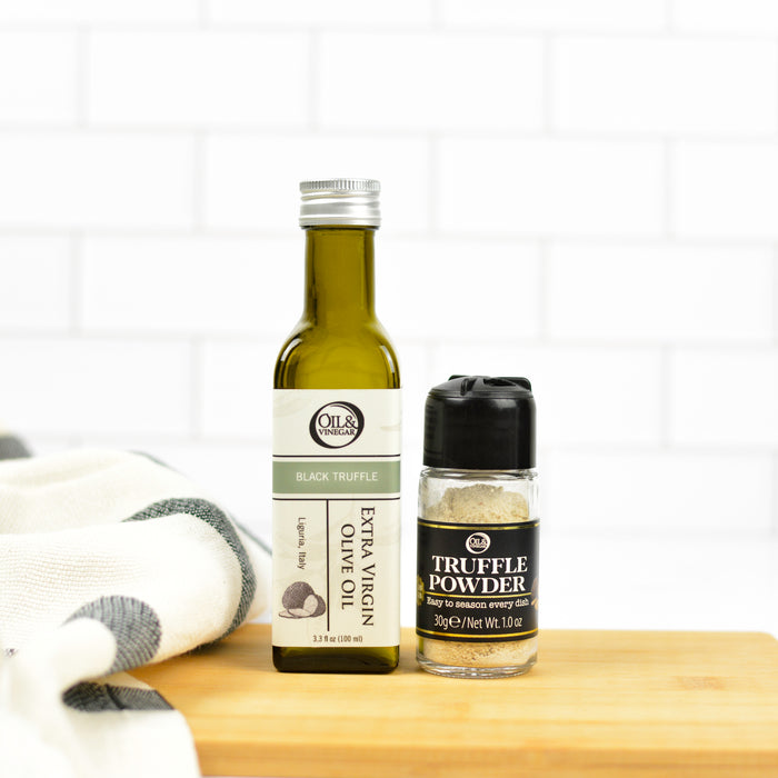 A bottle of black truffle extra virgin olive oil next to a shaker of truffle powder, placed on a wooden surface with a kitchen towel in the background.