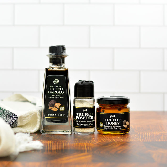 A set of truffle-related products including a bottle labeled 'Truffle Barolo', a smaller bottle with 'Truffle Powder', and a jar with 'Truffle Honey', arranged on a wooden surface with a white cloth in the background.