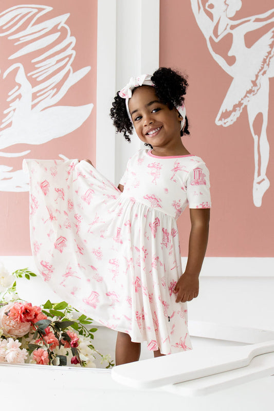 Young girl posing wearing the Twirl Dress in Toile Ballet standing in a white wooden rowboat beside flowers in front of ballet themed art