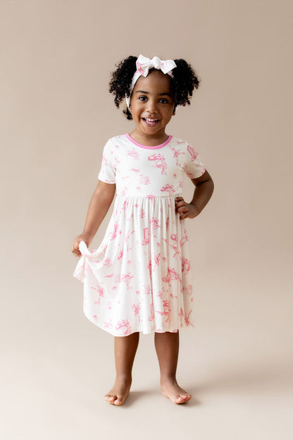 Smiling girl standing wearing the Twirl Dress in Toile Ballet with matching bow