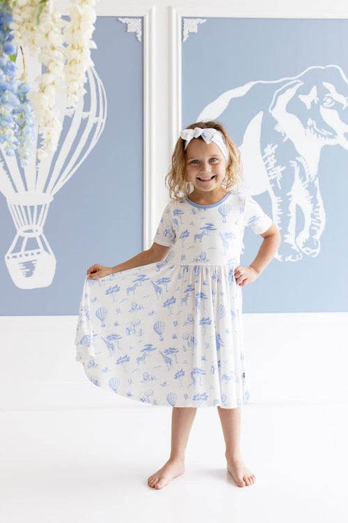 Young girl modeling the Twirl Dress in Toile Safari holding the skirt of the dress out standing in front of safari inspired artwork