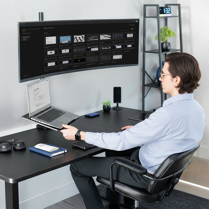 Person sitting at a desk with a large monitor and laptop, in a home office setting.