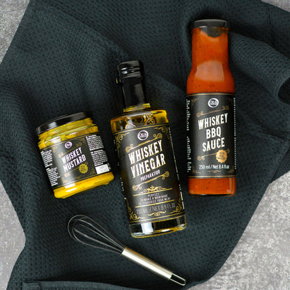 A set of whiskey-themed condiments including whiskey vinegar, whiskey BBQ sauce, and whiskey mustard, presented with a small whisk.