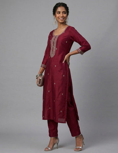 A woman modeling a deep maroon three-piece kurta set with silver embroidery on the neckline and cuffs, paired with matching trousers and silver high-heeled sandals, against a solid gray studio background.