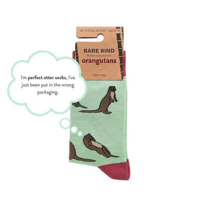 A pair of crew socks with a light green base and brown otter illustrations, attached to a cardboard hanger labeled 'BARE KIND'. A light blue speech bubble says, 'I'm perfect otter socks, I've just been put in the wrong packaging.