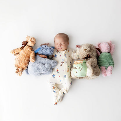 Sleeping newborn laying on a light neutral surface wearing the Zippered Footie in Winnie the Pooh Celebration surrounded by stuffed animals